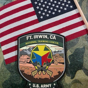May include: A patch for the U.S. Army's National Training Center at Ft. Irwin, CA, featuring crossed rifles, tanks, and a geometric design in yellow, blue, and red. An American flag is in the background.