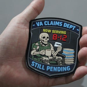 May include: A shield-shaped morale patch featuring a skeleton in camouflage clothing sitting in a chair with a coffee mug and a stack of files. The text reads VA CLAIMS DEPT, NOW SERVING 8-12, and STILL PENDING in blue, yellow, and red lettering.
