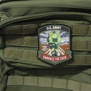 May include: An olive green tactical backpack featuring a rectangular morale patch. The patch displays a soldier with a large backpack walking on a path, with the text U.S. ARMY at the top and EMBRACE THE SUCK on a red banner at the bottom.