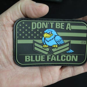 May include: A rectangular morale patch featuring an olive green and black American flag background. A blue falcon with yellow talons and beak sits atop two olive green military chevrons. The text reads DON'T BE A BLUE FALCON in olive green.