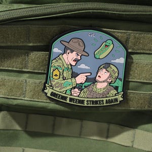 May include: A morale patch featuring a cartoon illustration of a drill sergeant yelling at a soldier, with a floating hot dog character above them. The text "GREENIE WEENIE STRIKES AGAIN" is printed on a banner at the bottom of the patch.