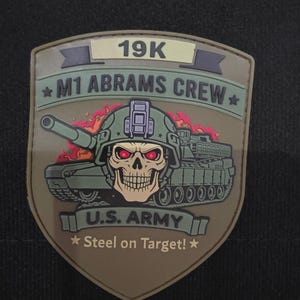 May include: A shield-shaped patch with the text "19K" and "M1 ABRAMS CREW". The patch features a tank illustration with a skull wearing a helmet. The text "U.S. ARMY" and "Steel on Target!" are also present. The patch has a tan and green color scheme.