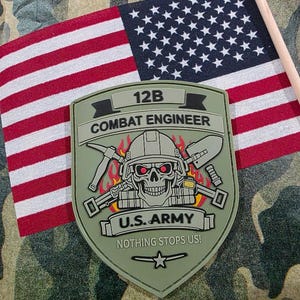 May include: An olive green 12B Combat Engineer patch with a skull wearing a helmet, crossed pickaxe and shovel, and the text "U.S. Army" and "Nothing Stops Us!" is displayed. An American flag is in the background.