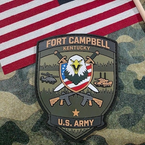 May include: A military patch with the text "FORT CAMPBELL KENTUCKY" and "U.S. ARMY". The patch features an eagle, crossed swords, rifles, and military vehicles. An American flag is in the background.