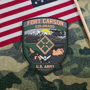 May include: A patch featuring the words "Fort Carson Colorado" with a military emblem. The emblem includes a tank, helicopters, a mountain scene, and the text "U.S. Army". An American flag is in the background.