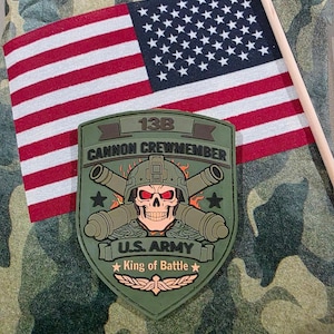 May include: A military patch with the text "13B Cannon Crewmember" and a skull wearing a helmet, with the words "U.S. Army" and "King of Battle". An American flag is in the background.