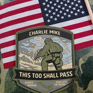 May include: A military-themed patch with the text "CHARLIE MIKE" at the top and "THIS TOO SHALL PASS" below. The patch features a soldier with a weapon and lightning in the background. An American flag is in the background.