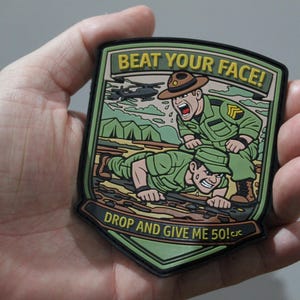 May include: A shield-shaped morale patch featuring a cartoon illustration of a drill sergeant yelling at a soldier doing push-ups in the mud. The text reads BEAT YOUR FACE! at the top and DROP AND GIVE ME 50! at the bottom in yellow lettering.