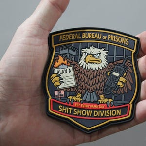 May include: A shield-shaped morale patch featuring a cartoon eagle holding a burning clipboard labeled PLAN A and a radio. The text reads FEDERAL BUREAU OF PRISONS, EST EVERY DAMN DAY, and SHIT SHOW DIVISION. A small sign says DO NOT RUN.