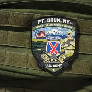 May include: A shield-shaped patch attached to an olive green tactical bag. The patch features the text FT. DRUM, NY, HOME OF THE 10TH MOUNTAIN DIVISION, and U.S. ARMY. It depicts a mountain, a soldier, a tank, an American flag, and crossed rifles.