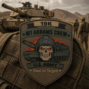 May include: A military patch featuring a skull wearing a helmet with red eyes, flames, and a tank graphic. Text includes "19K", "M1 ABRAMS CREW", "U.S. ARMY", and "Steel on Target!". A tank and soldiers are in the background.