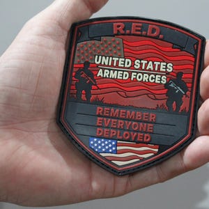 May include: A shield-shaped patch with a black border featuring the text R.E.D. at the top, UNITED STATES ARMED FORCES in the center, and REMEMBER EVERYONE DEPLOYED at the bottom. The design includes silhouettes of soldiers and American flags.