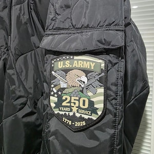 May include: Black quilted jacket with a patch featuring the U.S. Army emblem, an eagle, crossed rifles, and the text "250 YEARS SERVICE 1775-2025". The jacket has a pocket on the sleeve.