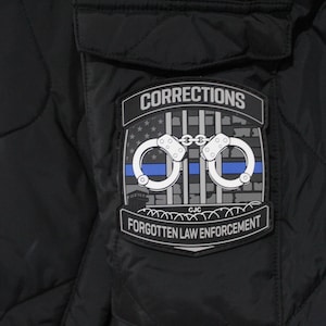 May include: A black quilted jacket featuring a rectangular patch with the text CORRECTIONS and FORGOTTEN LAW ENFORCEMENT. The patch displays a gray brick wall, prison bars, a blue horizontal line, and a pair of silver handcuffs in the center.