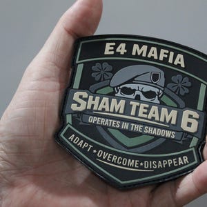 May include: A shield-shaped tactical morale patch in black, olive green, and tan. It features a skull wearing a beret, four-leaf clovers, and the text E4 MAFIA, SHAM TEAM 6, OPERATES IN THE SHADOWS, and ADAPT OVERCOME DISAPPEAR in raised lettering.