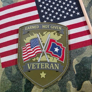 May include: An olive green Veteran patch with the text "EARNED - NOT GIVEN" and "VETERAN". The patch features crossed flags, one American and one with a blue star. An American flag is in the background.