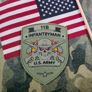 May include: An olive green 11B Infantryman patch with a skull graphic, crossed rifles, and the text "U.S. ARMY FOLLOW ME." An American flag is in the background, along with a camouflage pattern.