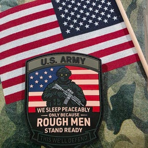 May include: A patch with the text "U.S. ARMY" and the words "WE SLEEP PEACEABLY ONLY BECAUSE ROUGH MEN STAND READY THIS WE'LL DEFEND" is displayed. An American flag is in the background. The patch features a soldier with a rifle.