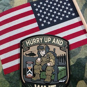 May include: A patch with the text "HURRY UP AND WAIT" featuring a soldier with a rifle, an hourglass, and a helicopter. An American flag is in the background. The patch is on a camouflage background.