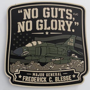 May include: A rectangular black and tan morale patch featuring a fighter jet with shark teeth nose art. The text reads "NO GUTS, NO GLORY." and "MAJOR GENERAL FREDERICK C. BLESSE" with a stylized American flag background and the tail number FG 829.