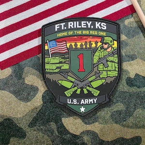 May include: A military patch with the text "FT. RILEY, KS HOME OF THE BIG RED ONE" and "U.S. ARMY". The patch features a shield design with a red "1" and a soldier graphic. An American flag is in the background.
