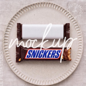 May include: Two rectangular candy bars placed horizontally on a textured plate. The top bar features a plain white wrapper, while the bottom bar is a Snickers candy bar with its signature brown, blue, and white branding and a visible peanut texture.
