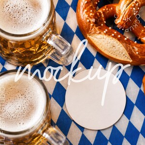 以下が含まれることがあります： 黄金色のビールと白い泡が入った2つのガラス製ビールジョッキ、大きな塩味のプレッツェル、そして青と白のひし形模様のテーブルクロスの上に置かれた空白の円形白いコースター。画像にはmockupと4Kというテキストが含まれています。