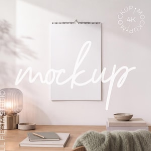 May include: A vertical wall calendar mockup with a white blank page and a black wire-o binding at the top. The calendar is centred on a light wall above a wooden desk featuring a ribbed glass lamp, a small notebook with a pen, and stacked books.