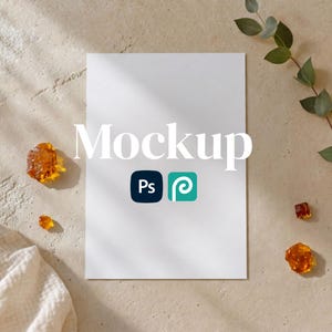Puede incluir: Una maqueta de papel blanco rectangular centrada sobre una superficie beige texturizada. La palabra Mockup está impresa en blanco, acompañada de iconos Ps azul oscuro y P verde azulado. Piezas de resina ámbar y una rama de eucalipto verde.