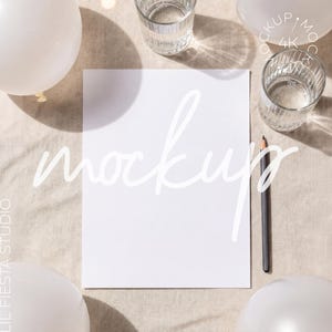 May include: A vertical white rectangular paper sheet centered on a neutral fabric surface, accompanied by a dark pencil, two clear ribbed drinking glasses, and three white balloons, with the word mockup written in white script across the center.