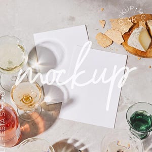 May include: Two blank white rectangular paper sheets positioned centrally, surrounded by several clear glass wine glasses, a bottle of rose wine, and a wooden board holding pieces of cheese and crackers. The text mockup 4K is visible.