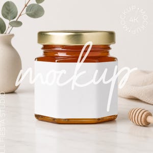 Pode incluir: Um frasco de vidro hexagonal cheio de mel de cor âmbar, com uma tampa de metal dourado e um rótulo retangular branco em branco na frente. O frasco está posicionado sobre uma superfície branca, acompanhado por um pau de mel de madeira.