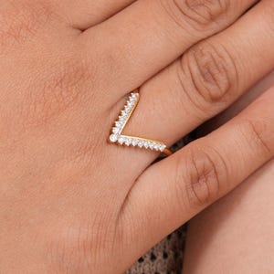 Könnte beinhalten: Ein goldfarbener, V-förmiger Ring mit kleinen, klaren Edelsteinen an den äußeren Kanten. Der Ring wird auf einer Hand mit einer neutral getönten Maniküre gezeigt.
