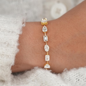 Puede incluir: Una pulsera dorada con una serie de piedras preciosas transparentes de diferentes formas. La pulsera presenta piedras de talla cuadrada, ovalada, pera y esmeralda. La pulsera se lleva en la muñeca, con un suéter blanco en el fondo.