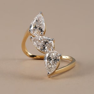 Puede incluir: Un anillo de oro con tres piedras transparentes en talla pera dispuestas en una línea diagonal escalonada. La banda es de oro amarillo pulido, mientras que las garras que sujetan cada una de las tres piedras facetadas son de metal plateado.