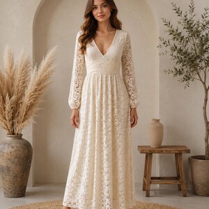 May include: A cream lace dress with long sleeves and a V-neck. The dress has a fitted waist and a full-length skirt. The model is wearing nude heels. The background includes a neutral-toned arch, a small wooden table, and decorative plants.