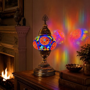 May include: A table lamp featuring a colorful mosaic glass shade with red, blue, and green geometric patterns. The lamp has an antique brass-toned metal base and a decorative finial on top, casting vibrant, multi-colored light reflections.