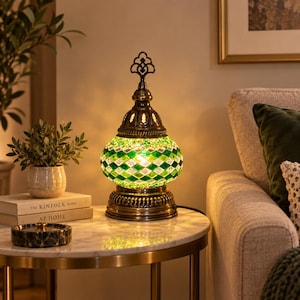 Può includere: Una lampada da tavolo con un paralume in vetro arrotondato caratterizzato da un motivo a mosaico a rombi verdi e bianchi. La lampada presenta una base in metallo color ottone antico e un puntale decorativo, emanando una luce calda.