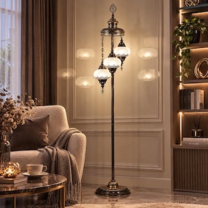 Op de afbeelding: Een hoge vloerlamp met een donkerbronzen metalen afwerking en drie hangende witte glazen bolvormige kappen. De lamp heeft gedetailleerde metalen filigraanversieringen rond de boven- en onderkant van elke kap, hangend aan een centrale paal op een ronde voet.