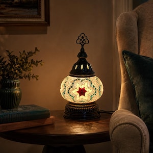 May include: A decorative table lamp with a mosaic glass shade. The lamp features a starburst design in red, green, and white. The base and top are black, and the lamp is plugged into an outlet. The lamp is on a wooden table.