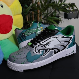 May include: Custom sneakers featuring a gray, teal, and white design with an eagle graphic. The shoes have black laces, green accents, and a white sole. The word "AIR" is visible on the side.