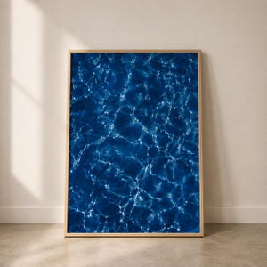 May include: A rectangular wall art print featuring a high-contrast photograph of deep blue water with bright white light reflections creating a rippling caustic pattern across the surface, encased in a thin, light-toned wooden frame.