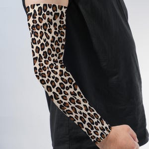 May include: A compression arm sleeve featuring a classic leopard print pattern with black and brown spots on a cream-colored background. The sleeve covers the forearm and upper arm, extending from the wrist to the bicep area with a fitted design.