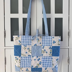 May include: A quilted tote bag featuring a patchwork design of blue and white gingham squares, solid light blue squares, and a white fabric pattern with blue blueberries and green leaves. The bag has two light blue striped shoulder straps.