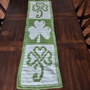 Puede incluir: Un camino de mesa de ganchillo con un diseño verde y blanco con tres tréboles. El camino de mesa tiene un borde verde.