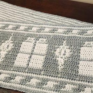 Könnte beinhalten: Ein grau-weißer Häkel-Schal mit einem sich wiederholenden Muster eines Raumschiffs. Der Schal hat Fransen an den Enden.