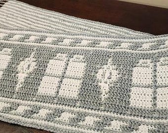 May include: A gray and white crocheted scarf with a repeating pattern of a spaceship. The scarf has fringe on the ends.