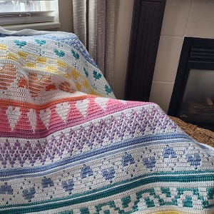 Puede incluir: Una manta de ganchillo colorida con un patrón de arcoíris de corazones, flores y formas geométricas. La manta está hecha con una variedad de colores de hilo, incluyendo amarillo, naranja, rojo, rosa, morado, azul, verde y blanco.