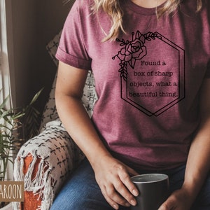 May include: A heather maroon t-shirt with a black graphic design featuring a floral illustration and the text "Found a box of sharp objects, what a beautiful thing."