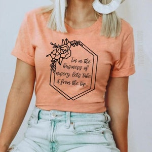 May include: A peach colored t-shirt with a black graphic print of a floral design and the text "I'm in the business of misery, let's take it from the top."